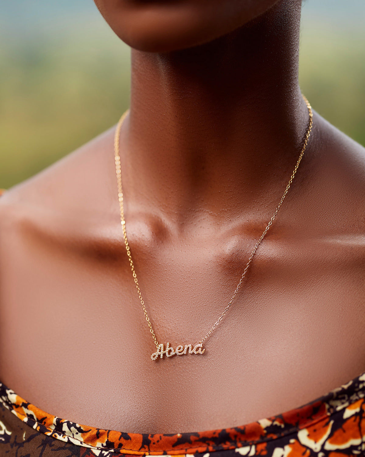 A close-up of a model wearing CASA London's 18k gold Abena Pavé Necklace. The delicate gold chain features a pendant with the name "Abena" intricately encrusted with shimmering pavé-set stones, celebrating the tradition of those born on a Tuesday. The model wears a patterned garment, providing a sophisticated backdrop that highlights the brand's commitment to elegant, narrative-driven luxury.