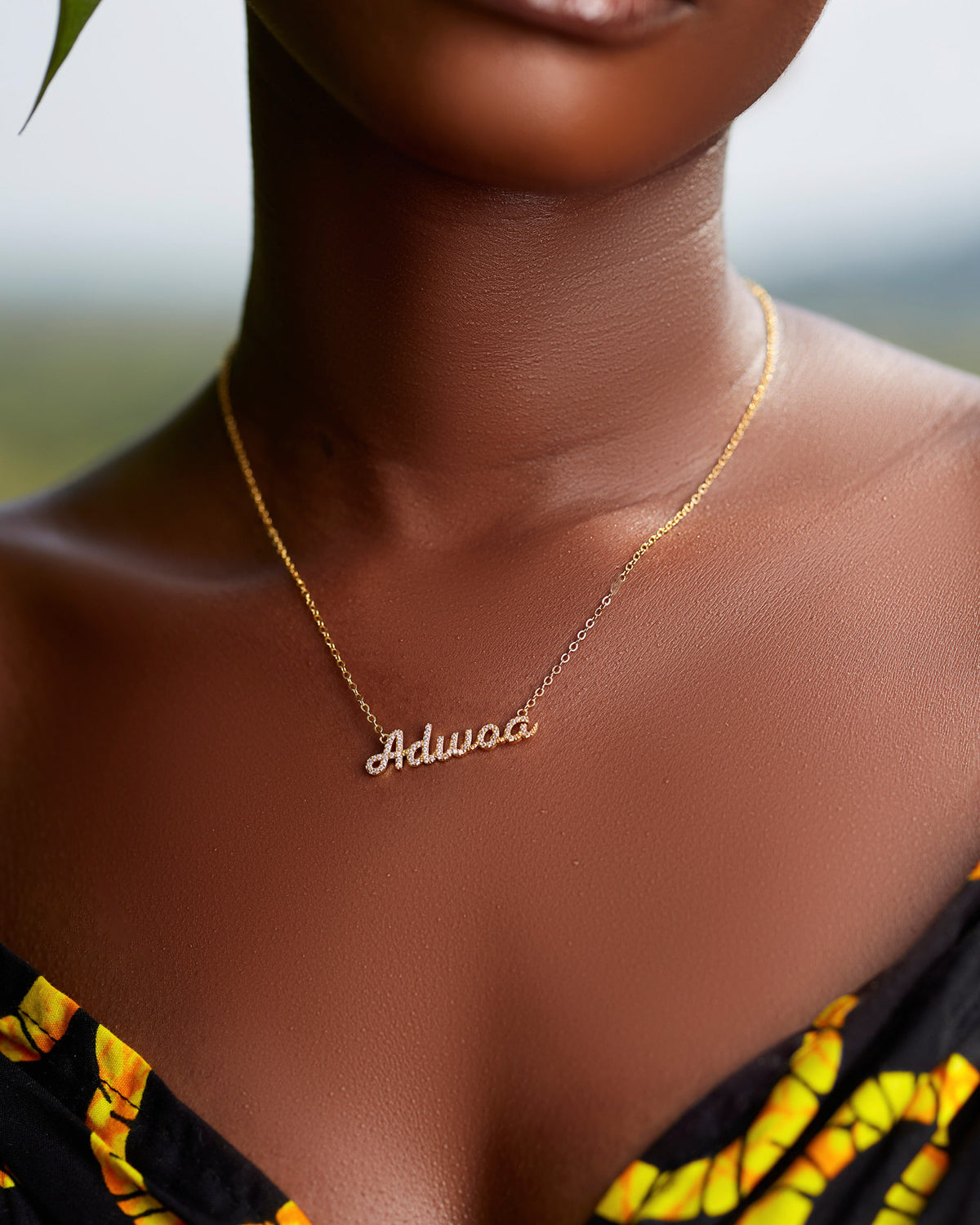 A close-up of a model wearing CASA London's 18k gold Adwoa Pavé Necklace. The delicate gold chain features a pendant with the name "Adwoa" intricately encrusted with shimmering pavé-set stones, celebrating the tradition of those born on a Monday. The model wears a black garment with a vibrant yellow pattern, providing a bold backdrop that highlights the brand's commitment to elegant, narrative-driven luxury.