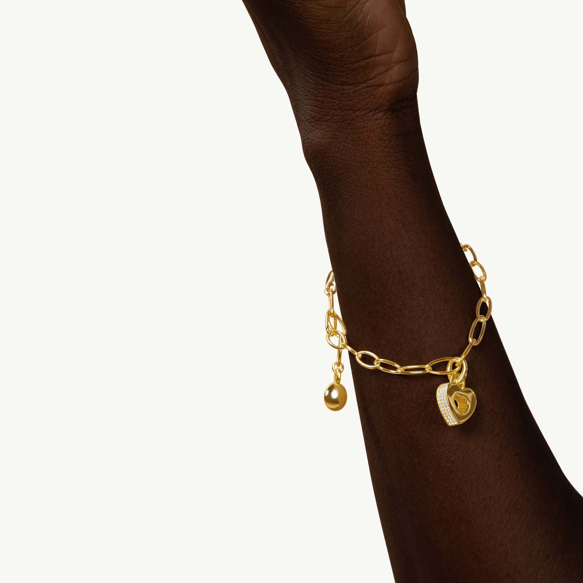 CASA LONDON Akoma Pavé Charm in 18ct gold vermeil worn on a dark-toned wrist. Heart-shaped Adinkra-inspired charm with white zirconia pavé stones on a gold link bracelet.