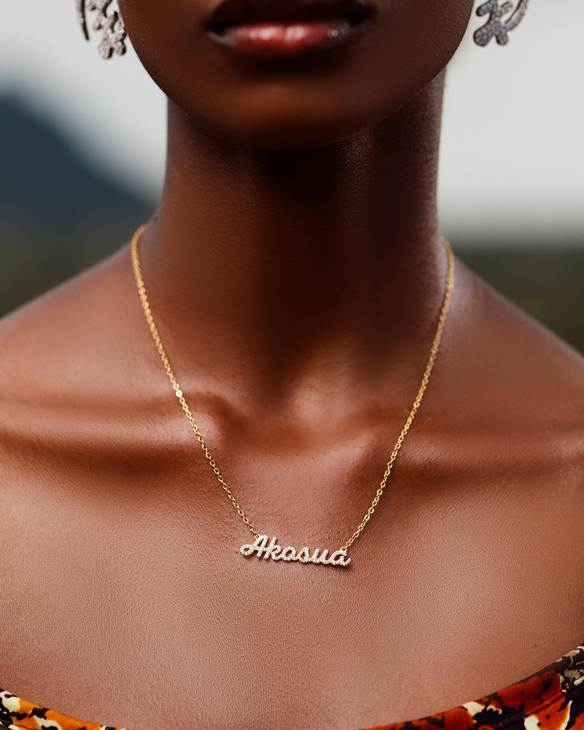 A close-up of a model wearing CASA London's 18k gold Akosua Pavé Necklace. The delicate gold chain features a pendant with the name "Akosua" intricately encrusted with shimmering pavé-set stones, celebrating the tradition of those born on a Sunday. The model wears a patterned garment, providing a sophisticated backdrop that highlights the brand's commitment to elegant, narrative-driven luxury.