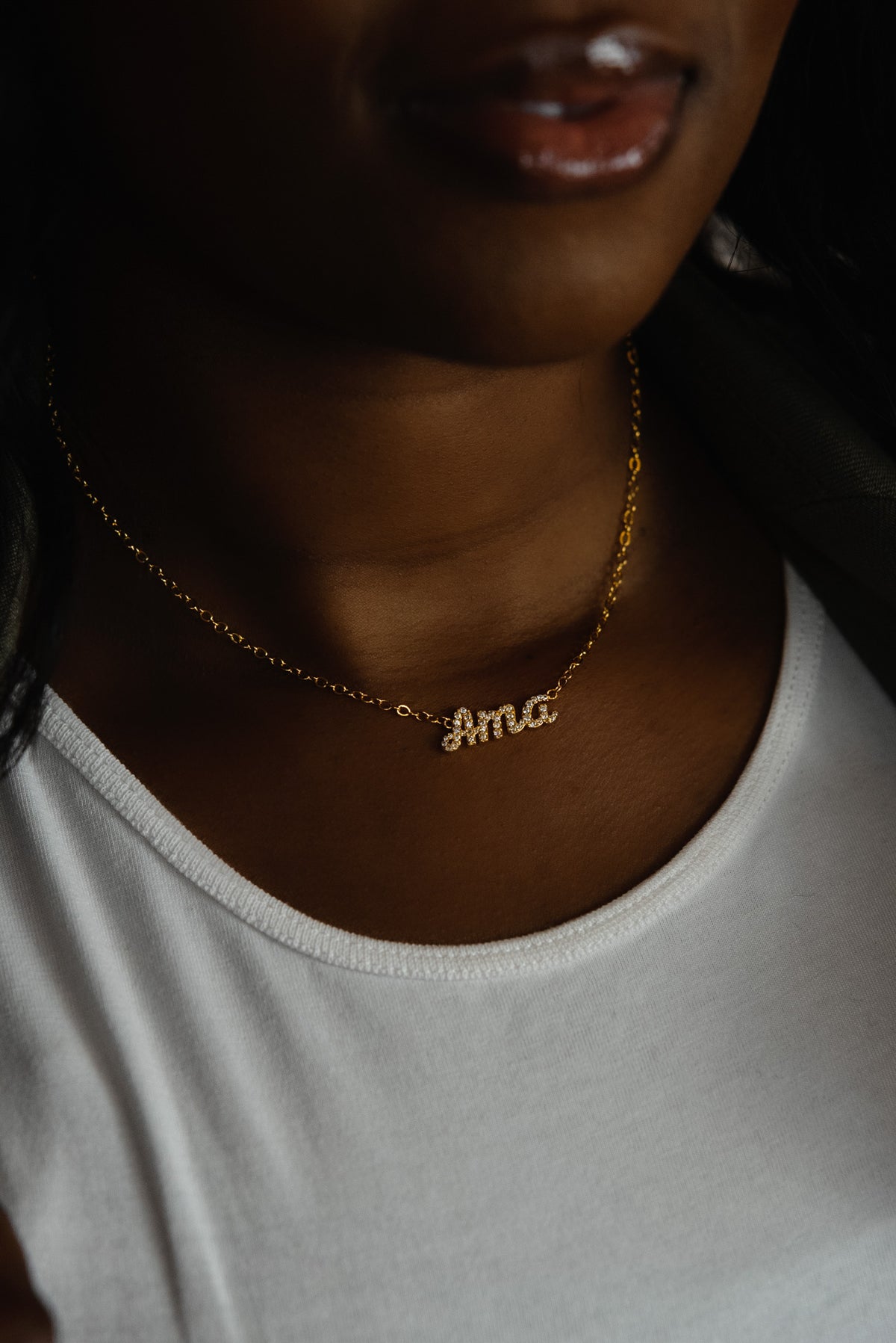 A close-up of a model wearing CASA London's 18k gold Ama Pavé Necklace. The delicate gold chain features a pendant with the name "Ama" intricately encrusted with shimmering pavé-set stones, celebrating the tradition of those born on a Saturday. The model wears a white top, providing a clean backdrop that highlights the brand's commitment to elegant, narrative-driven luxury.