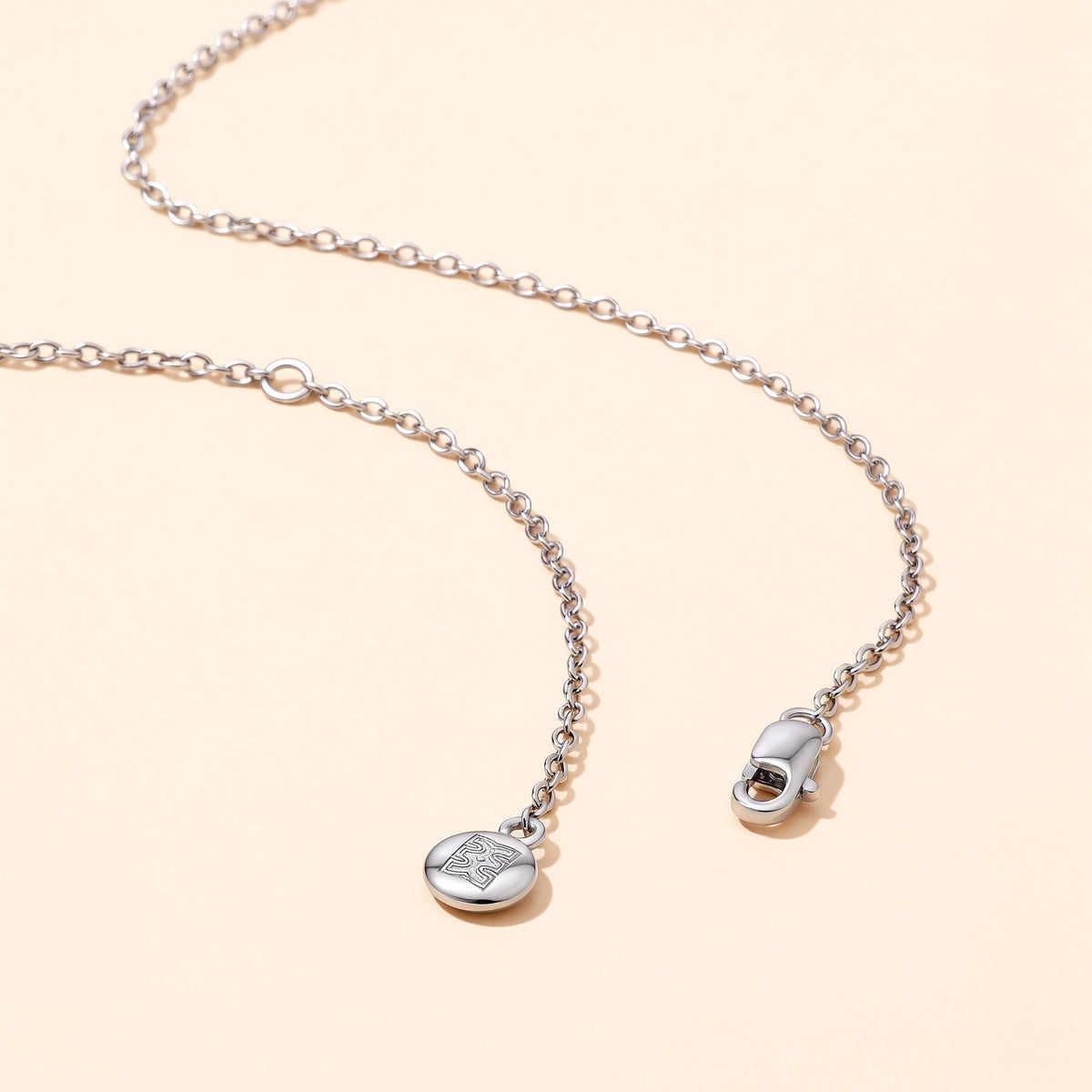 Close-up flat lay of the CASA London Custom Pavé Name Necklace chain in rhodium silver on S925 sterling silver, showing the lobster clasp and engraved CASA London brand insignia disc charm on a cream background