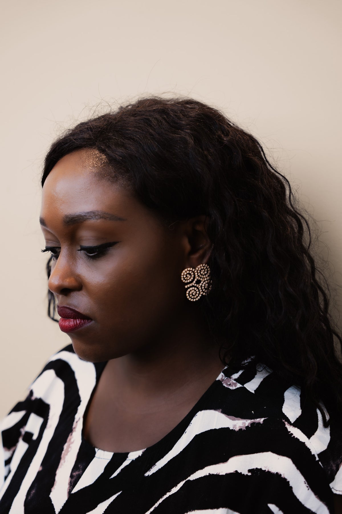 A close-up profile shot of a model wearing CASA London's gold Dwennimmen Pavé Earrings. The intricate earrings are shaped into the Adinkra symbol of rams' horns, representing strength and humility, and are meticulously encrusted with shimmering pavé-set stones. The detailed shimmer against the soft-focus backdrop highlights the brand's commitment to elegant, narrative-driven luxury.