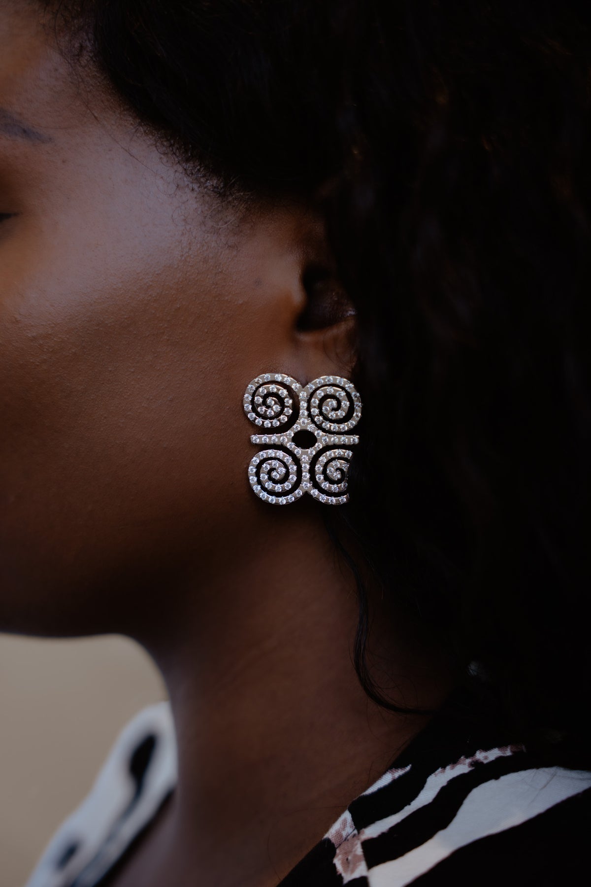 A close-up profile shot of a model wearing CASA London's silver Dwennimmen Pavé Earrings. The intricate earrings are shaped into the Adinkra symbol of rams' horns, representing strength and humility, and are meticulously encrusted with shimmering pavé-set stones. The detailed shimmer against the soft-focus backdrop highlights the brand's commitment to elegant, narrative-driven luxury.