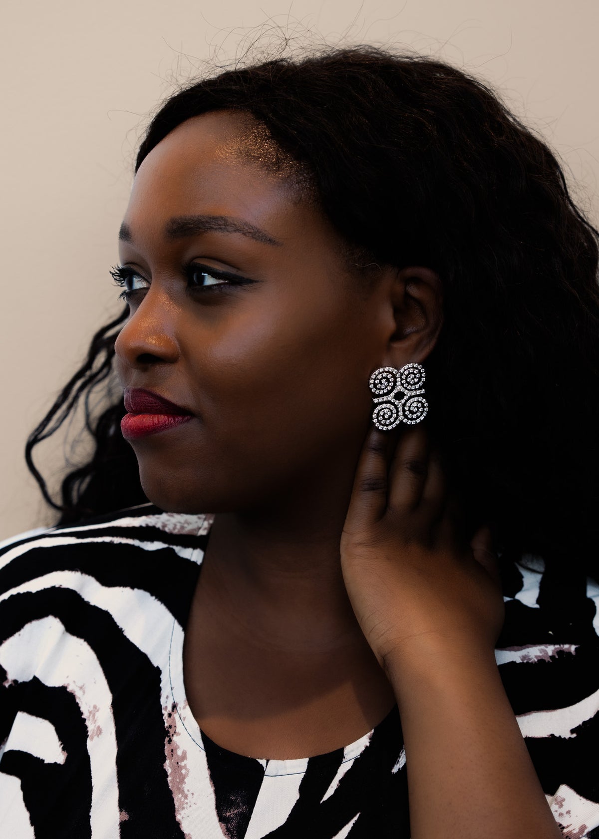 A close-up profile shot of a model wearing CASA London's silver Dwennimmen Pavé Earrings. The intricate earrings are shaped into the Adinkra symbol of rams' horns, representing strength and humility, and are meticulously encrusted with shimmering pavé-set stones. The detailed shimmer against the soft-focus backdrop highlights the brand's commitment to elegant, narrative-driven luxury.