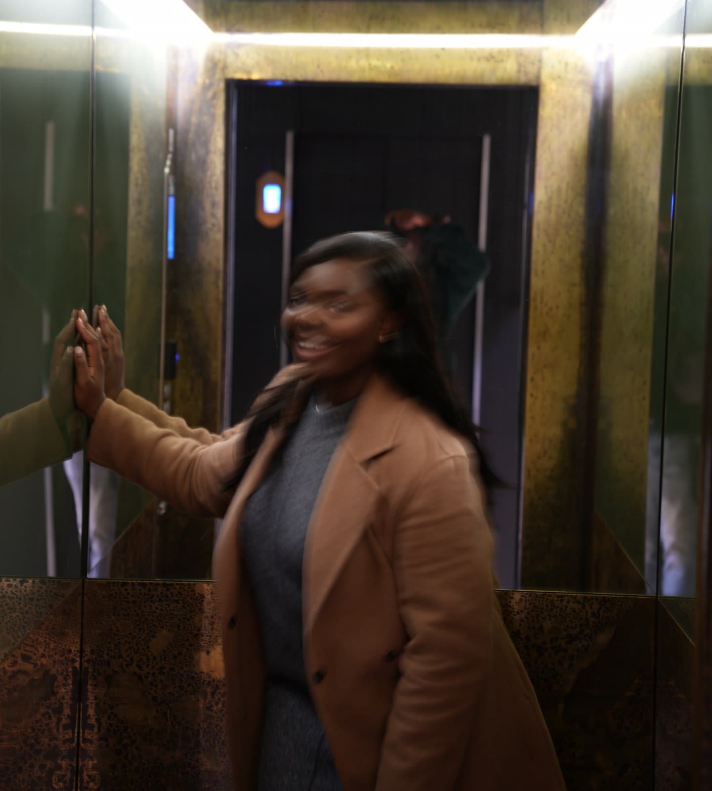 Christine, founder of CASA London, laughing and turning in motion with one hand against the lift mirror, wearing a camel coat