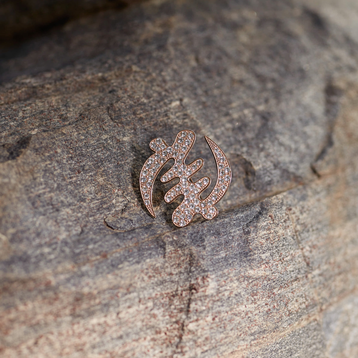 A detailed shot of CASA London's 18ct rose gold plated Gye Nyame lapel pin resting on the textured surface of a grey rock. The pin, meticulously shaped into the iconic Adinkra symbol and encrusted with shimmering pavé-set stones, catches the soft natural light. This outdoor setting highlights the brand's connection to heritage and its commitment to merging cultural significance with high-end luxury.