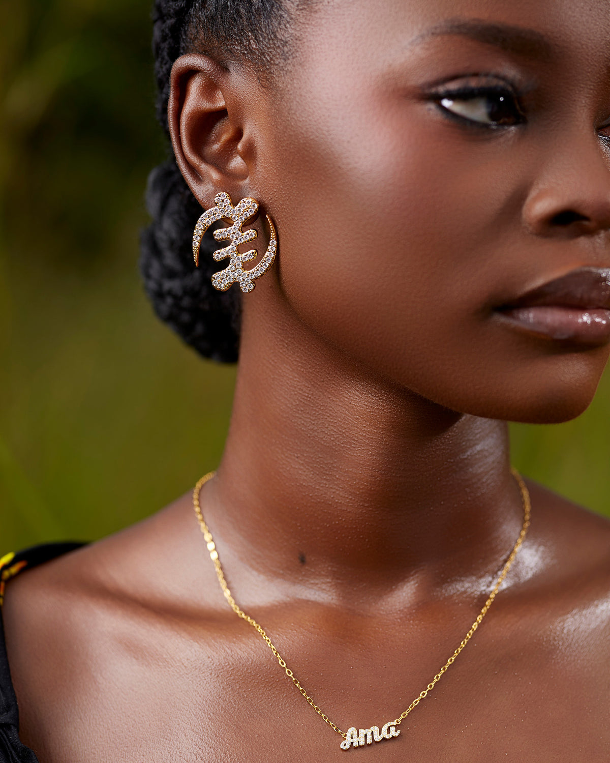 Close-up of a model wearing a large gold Gye Nyame Adinkra symbol earring, featuring a bold pavé-set high-grade zircon design against a natural outdoor background.
