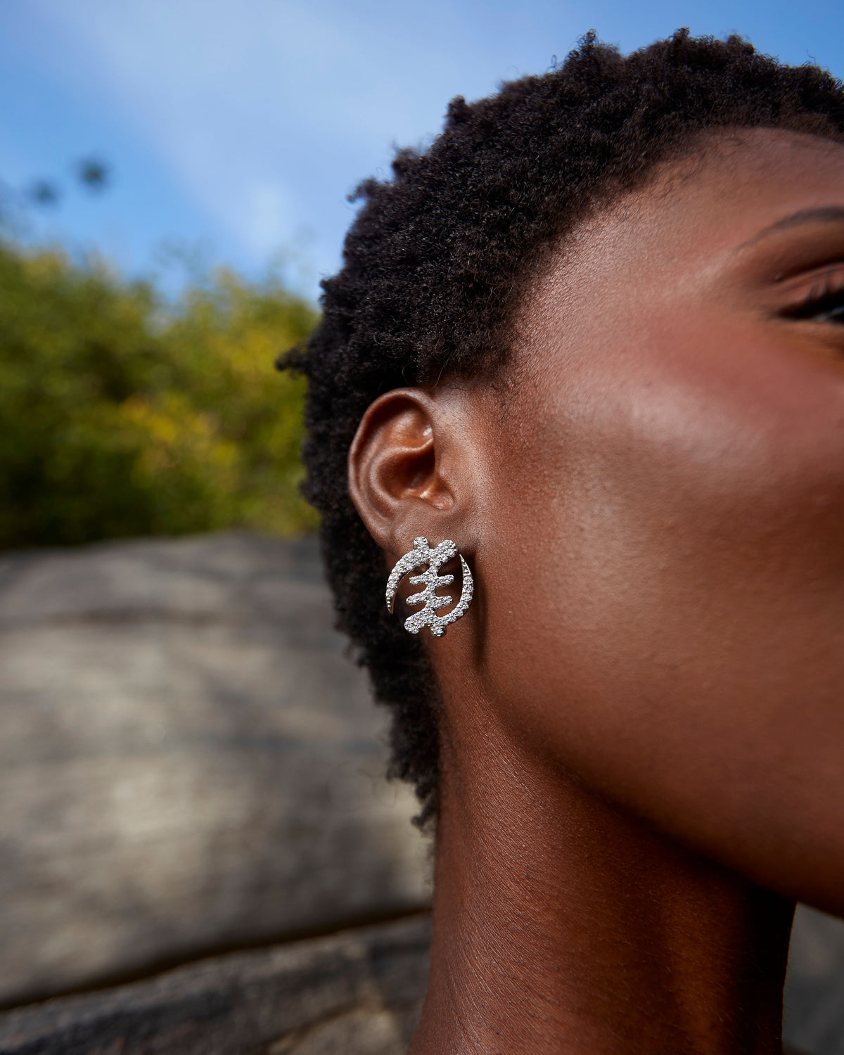 A close-up profile of a model wearing CASA London's silver Gye Nyame Pavé Earrings small. The earrings are intricately shaped into the iconic Adinkra symbol, meticulously encrusted with shimmering pavé-set stones that catch the natural outdoor light. The model’s natural hair and the soft focus of the rocky background highlight the brand's commitment to blending cultural heritage with contemporary luxury.
