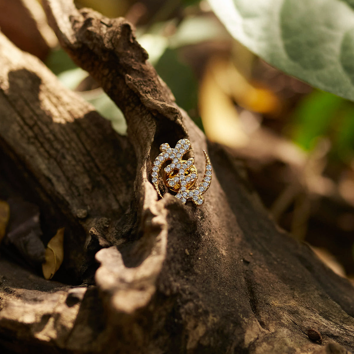 A detailed shot of CASA London's 18k gold plated Gye Nyame lapel pin nestled within the textured bark of a tree. The pin, intricately shaped into the iconic Adinkra symbol and encrusted with shimmering pavé-set stones, is highlighted by warm, natural sunlight. This outdoor setting emphasizes the brand's connection to heritage and its commitment to merging cultural significance with high-end luxury.