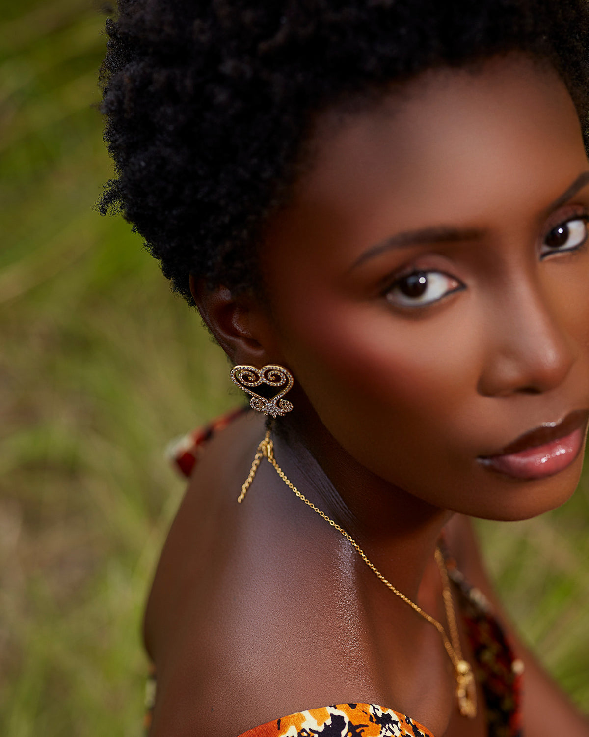 A close-up of a model wearing CASA London's gold Sankofa Pavé earrings. The earrings feature an intricate, heart-shaped design inspired by the Sankofa Adinkra symbol, encrusted with shimmering pavé-set stones. The model’s natural hair and the soft, natural light of the outdoor setting highlight the brand's commitment to blending cultural heritage with contemporary luxury.
