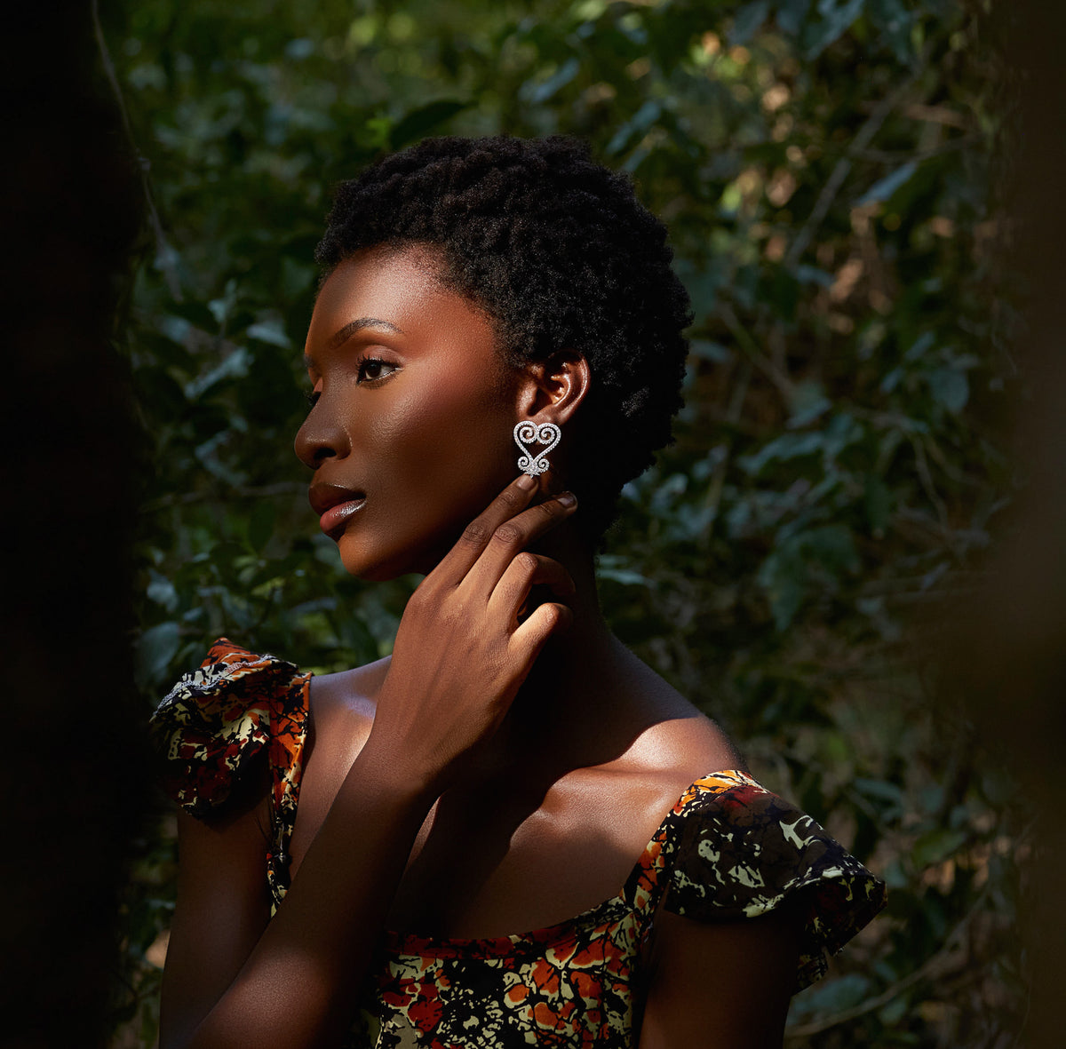 A portrait of a model wearing CASA London's silver Sankofa Pavé earrings. The earrings feature an intricate, heart-shaped design inspired by the Sankofa Adinkra symbol, encrusted with shimmering pavé-set stones. The model's natural hair and the soft, dappled sunlight of the lush outdoor setting highlight the brand's commitment to blending cultural heritage with contemporary luxury.