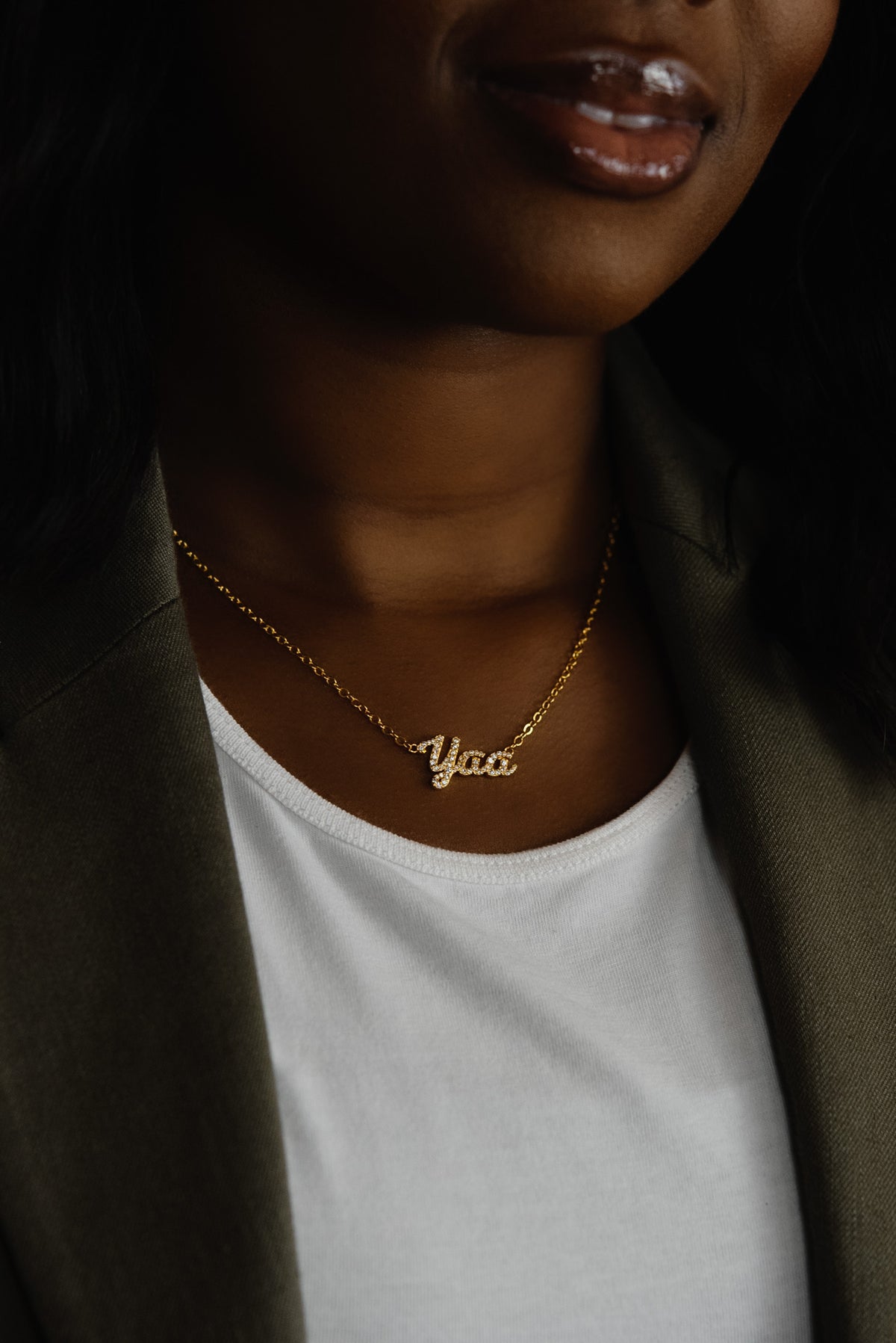 A close-up of a model wearing CASA London's 18k gold Yaa Pavé Necklace. The delicate gold chain features a pendant with the name "Yaa" intricately encrusted with shimmering pavé-set stones, celebrating the tradition of those born on a Thursday. The model wears a white top layered under an olive-green blazer, providing a sophisticated backdrop that highlights the brand's commitment to elegant, narrative-driven luxury.