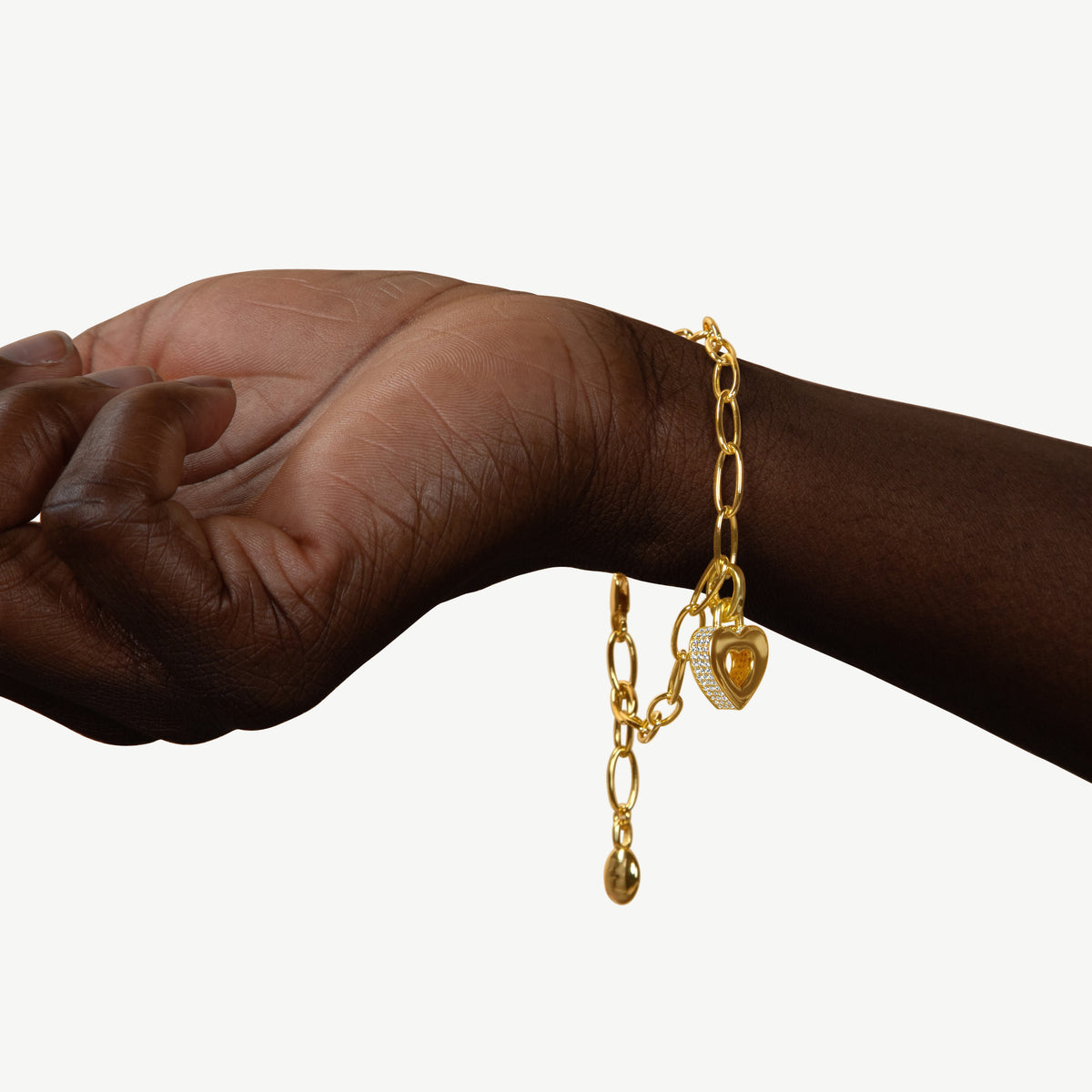CASA LONDON Akoma Pavé Charm bracelet in 18ct gold vermeil worn on a dark-toned hand and wrist. Chunky oval link chain featuring a heart-shaped zirconia pavé charm with an open Adinkra heart motif and CASA LONDON branded gold tag.